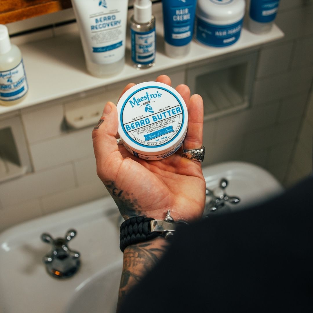 Tattooed hand holding Maestro’s Classic Mark of a Man Beard Butter jar beside other Maestro’s products in a modern bathroom setting.