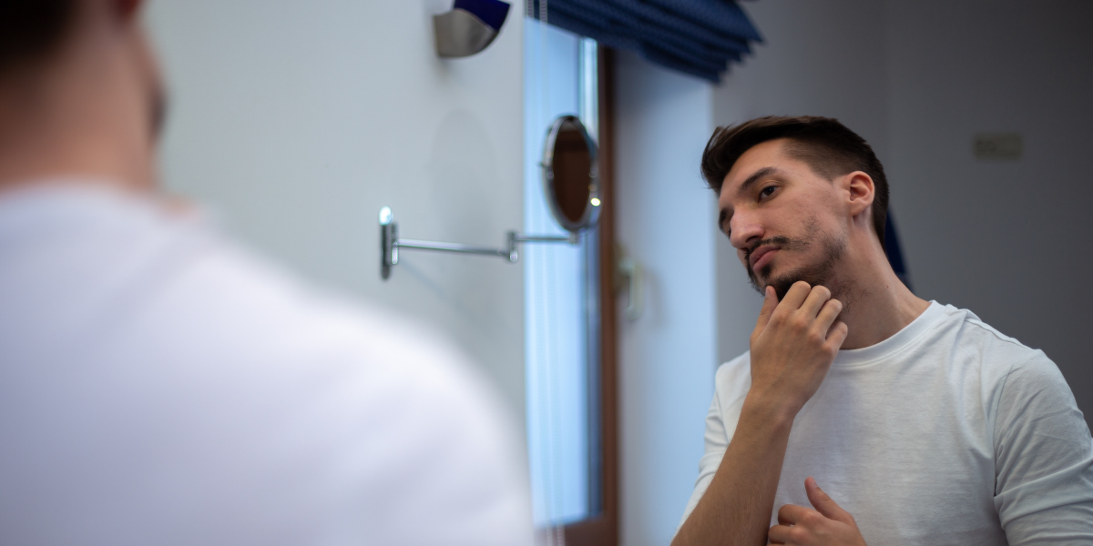man looking in mirror at patchy beard wondering what to do