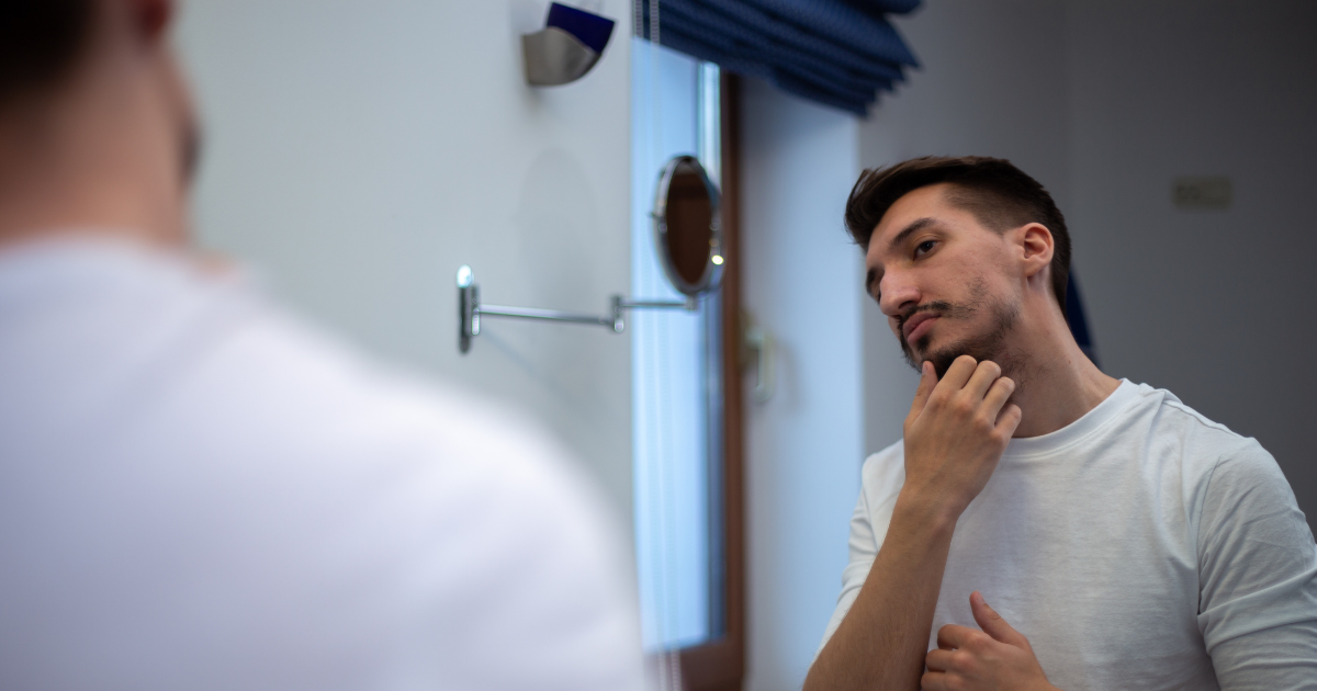 man looking in mirror at patchy beard wondering what to do