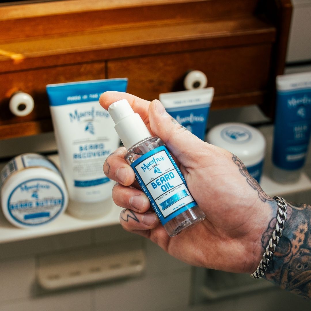 Tattooed hand holding Maestro’s Classic Mark of a Man Beard Oil bottle beside other beard care products on a bathroom counter.