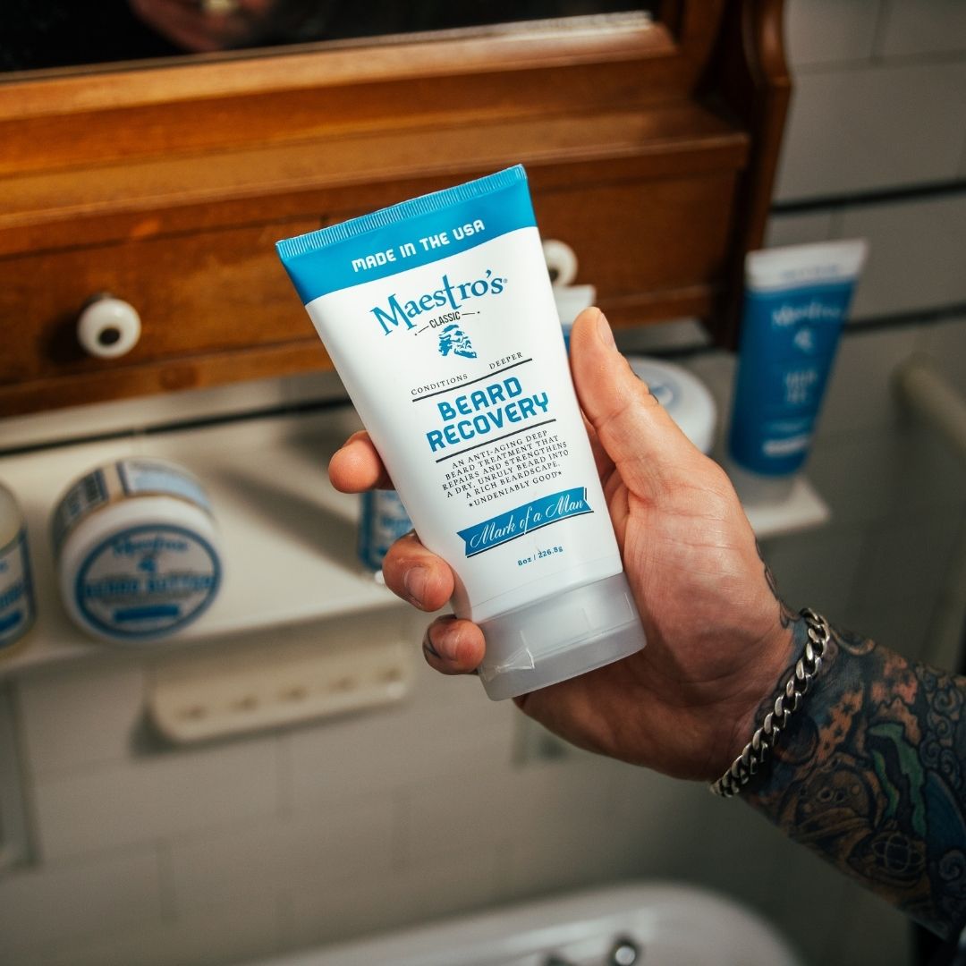 Tattooed hand holding Maestro’s Classic Mark of a Man Beard Recovery tube beside other Maestro’s products on bathroom counter.